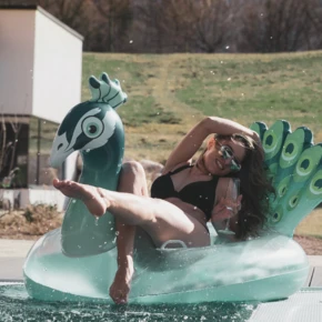 Frau entspannt sich auf einem Pfauen-Schwimmring am Pool, mit einem Drink in der Hand, umgeben von grüner Landschaft.