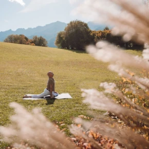 Eine Person meditiert auf einer Matte in einer grünen Wiese, umgeben von Bergen, ideal für Wellness und Entspannung im Hotel Klosterhof.