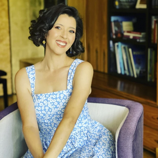 Eine lächelnde Frau in einem blauen Blumenkleid sitzt entspannt in einem Sessel, umgeben von Bücherregalen.