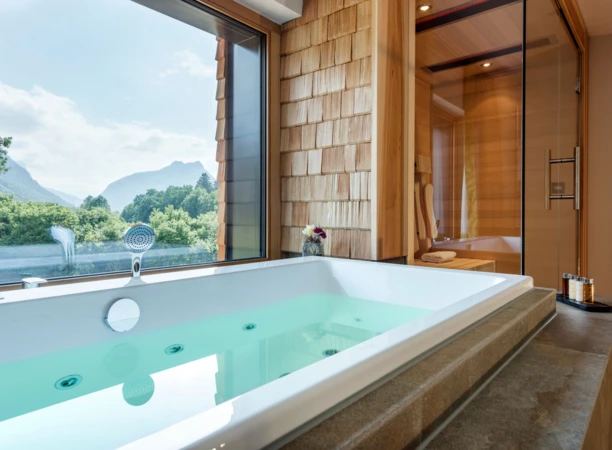 Blick auf eine Badewanne mit grünem Wasser und Aussicht auf Berge durch große Fenster in einem modernen Wellnessbereich.