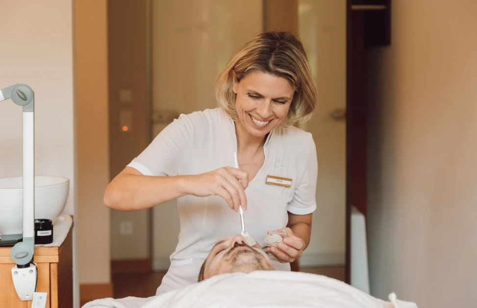 Eine Therapeutin trägt in einem Spa des Hotel Klosterhof eine Gesichtsmaske auf einen entspannten Gast auf.