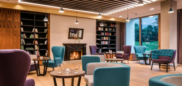 Elegant lounge with cozy seating, a fireplace, and bookshelves, offering a relaxing atmosphere. Large window with a view of greenery outside.