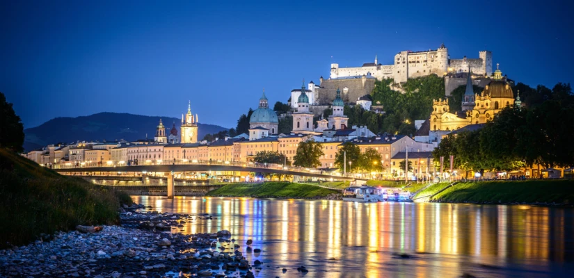 Salzburg bei Nacht mit beleuchteter Altstadt und Festung, reflektiert im Wasser des Flusses. Malerische Stimmung und historische Architektur.