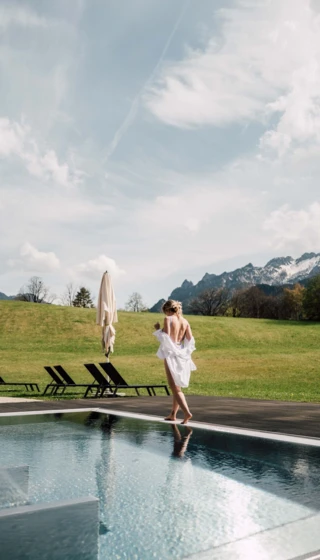 Eine Person in einem weißen Bademantel geht entspannt am Pool entlang, umgeben von einer grünen Landschaft und Bergen im Hintergrund.