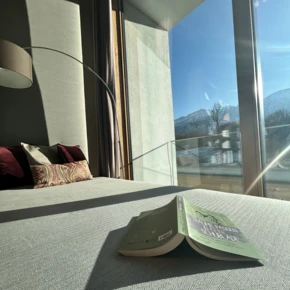 Ein gemütliches Zimmer im Hotel Klosterhof mit Blick auf die Berge und einem offenen Buch auf dem Bett. Viel Licht strömt durch das Fenster.