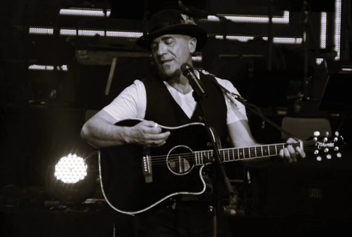 Musician playing acoustic guitar on stage, wearing a hat and vest, with stage lights in the background.