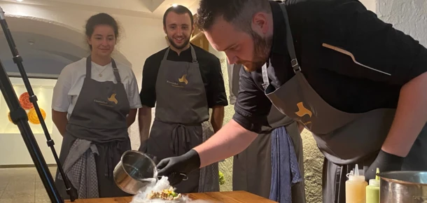 Ein Koch serviert ein Gericht mit dampfendem Nebel, während zwei Assistenten aufmerksam zuschauen. Holztafel und Küchengeräte sind sichtbar.