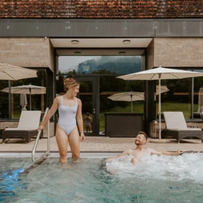 Eine Frau in einem wei&szlig;en Badeanzug steht am Poolrand, w&auml;hrend ein Mann im Wasser entspannt. Hintergrund zeigt das Hotel Klosterhof in Bayern.