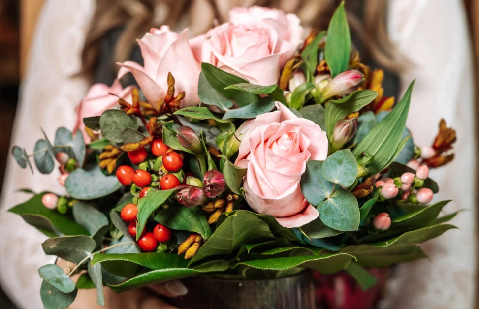 Eine Frau hält einen Strauß mit rosa Rosen und buntem Grün in einer Vase, lächelnd vor einem Holz-Hintergrund.