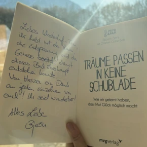 Eine Person hält ein Buch mit der Aufschrift "Träume passen in keine Schublade" vor einer Fensteransicht auf die Berge.