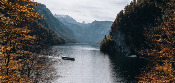 Ein Boot fährt auf dem ruhigen Wasser eines Bergsees, umgeben von herbstlichen Bäumen und majestätischen Bergen im Hintergrund.