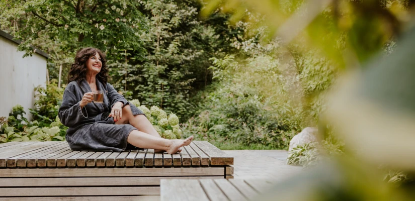 Frau in einem Bademantel sitzt entspannt auf einer Holzliege und genießt eine Tasse Tee in einer grünen Gartenlandschaft.