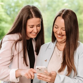 Zwei Frauen lächeln und schauen gemeinsam auf ein Smartphone, umgeben von natürlichem Licht und grüner Landschaft.