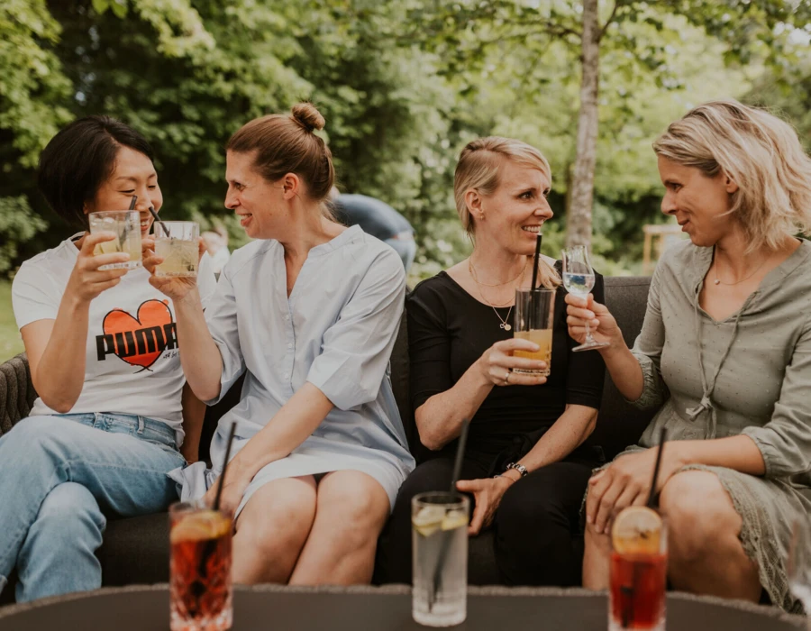 Fünf Frauen genießen Drinks auf einer Couch im Freien, umgeben von grünem Ambiente. Freundliche Stimmung und geselliges Beisammensein.