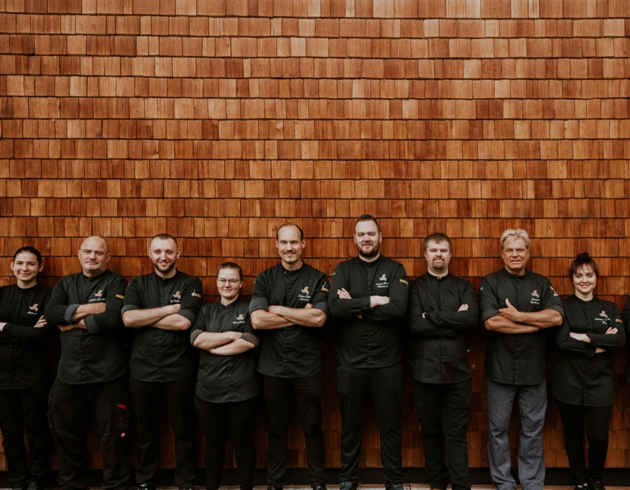 Eine Gruppe von Köchen in schwarzen Uniformen, die stolz vor einer Holzverkleidung stehen, lächeln und posieren zusammen.