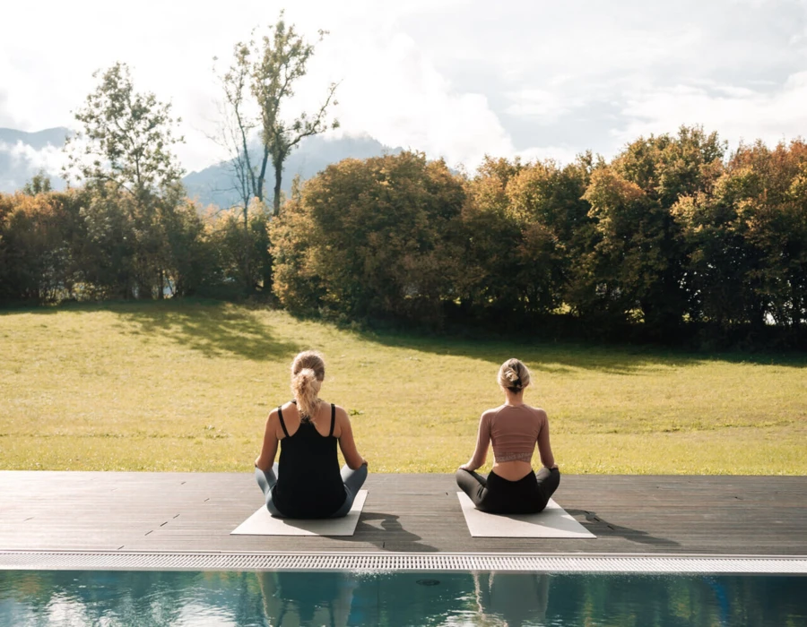 Two women practicing yoga by a pool at Wellnesshotel Klosterhof in Bayern, surrounded by lush greenery and a serene landscape.