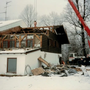Ein Kran hebt Teile des Daches eines Hauses ab, während Arbeiter im Schnee an der Baustelle tätig sind.