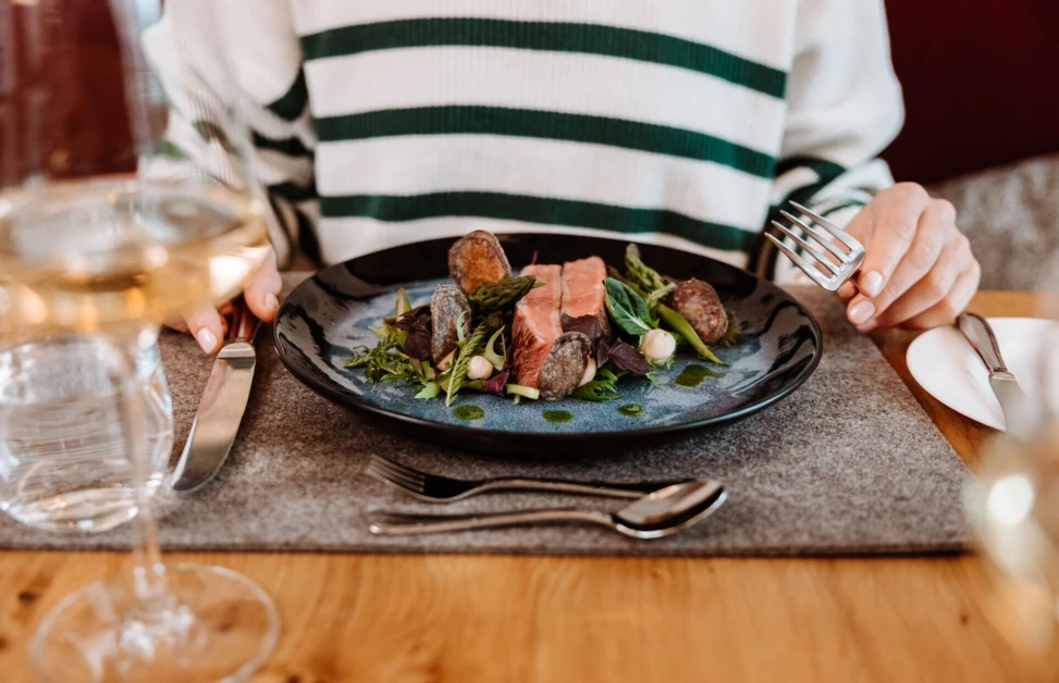 A person dining with a gourmet dish of meat, vegetables, and herbs on a plate, accompanied by a glass of white wine.