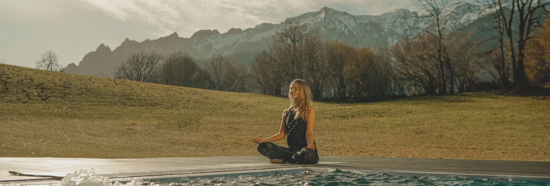 A woman meditating outdoors near a bubbling pool with mountains in the background.