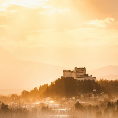 Die Burg auf einem Hügel, umgeben von sanften Hügeln und Nebel, beleuchtet von goldenem Sonnenlicht.