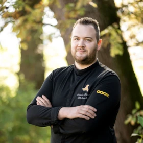 Chef in black uniform standing outdoors with arms crossed, surrounded by greenery.