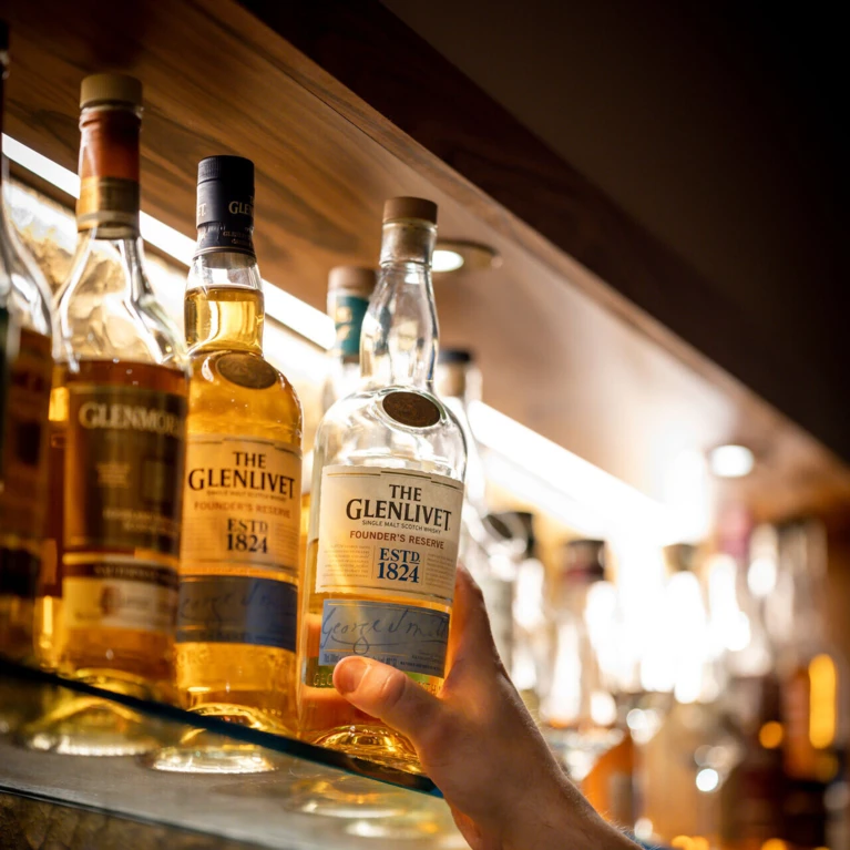 A hand reaches for a bottle of Glenlivet whisky on a well-stocked bar shelf at Klosterhof wellness hotel.