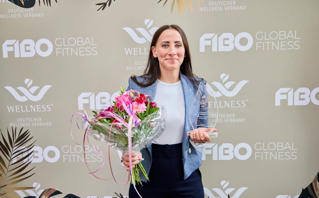 Eine Frau h&auml;lt eine Auszeichnung und einen Blumenstrau&szlig; vor einem FIBO Global Fitness Hintergrund.