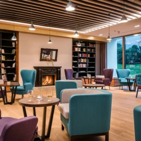 Gemütlicher Lounge-Bereich im Hotel Klosterhof mit Leserecke, bequemen Sitzgelegenheiten und Kamin, umgeben von natürlichem Licht.