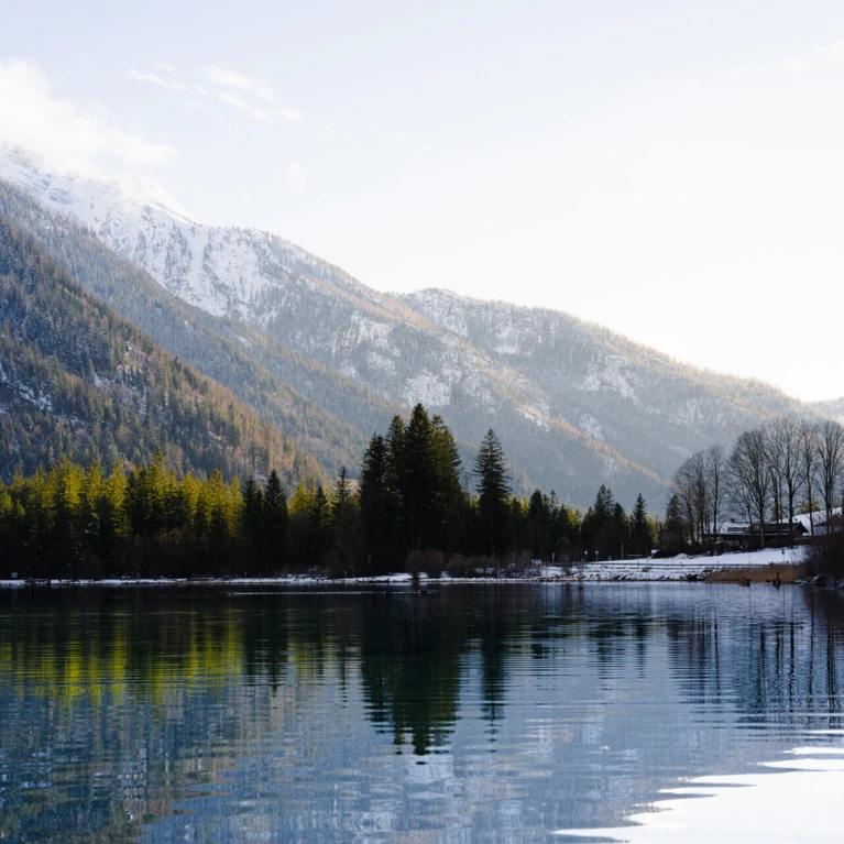 Bergsee mit klarer Wasseroberfläche, umgeben von schneebedeckten Bergen und dichten Wäldern unter blauem Himmel.