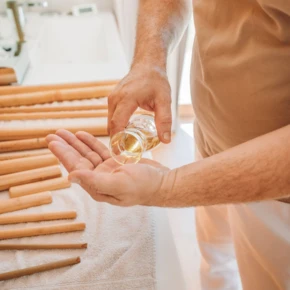 Ein Masseur gießt Öl in seine Hand, bereit für eine entspannende Massage, umgeben von Massagestäben auf einem Handtuch.
