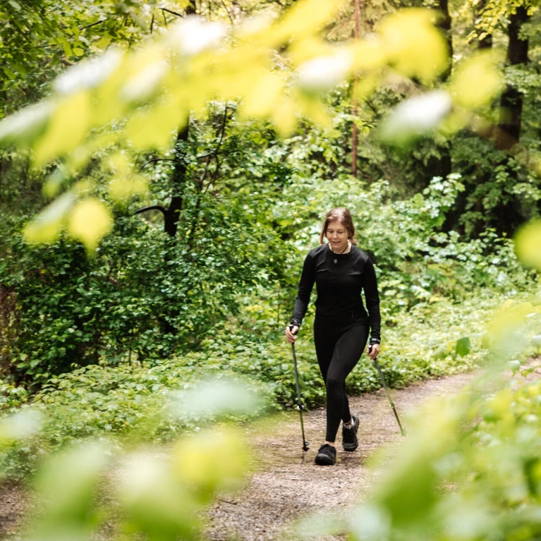 Eine Frau wandert mit Stöcken auf einem Waldweg, umgeben von üppigem Grün und Bäumen.