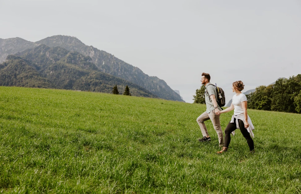 Ein Paar wandert Hand in Hand über eine grüne Wiese mit Bergen im Hintergrund.