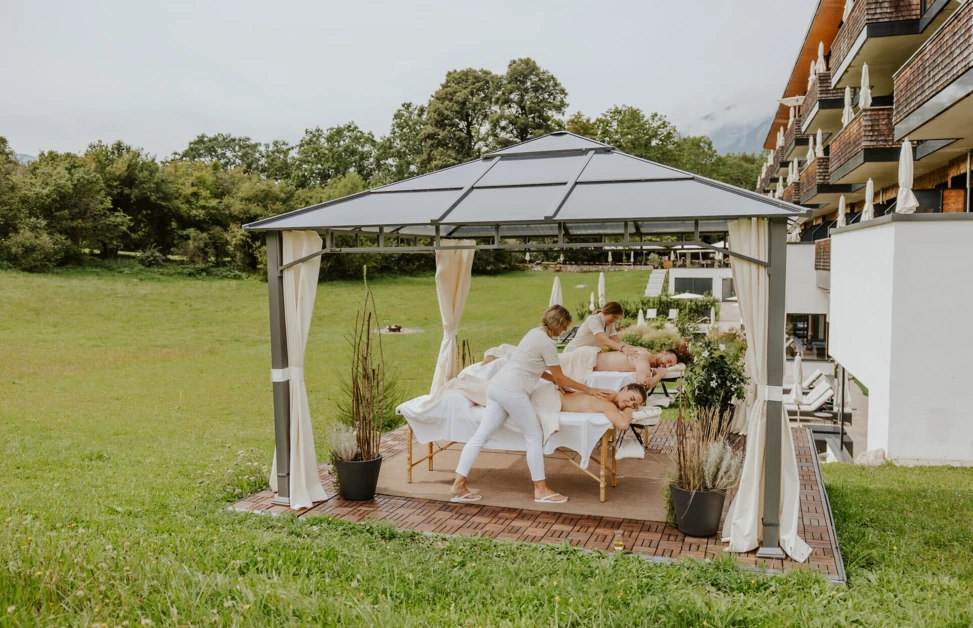 Outdoor massage area with therapists providing treatments under a gazebo, surrounded by greenery and near a building.