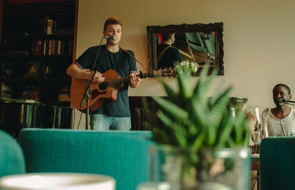 Musician in a cozy café setting at Wellnesshotel Klosterhof in Bayern, playing guitar near teal chairs, with a plant and candle in the foreground.