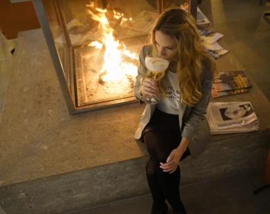 Eine Frau sitzt neben einem Kamin und genießt einen Cocktail in einem modernen, stilvollen Ambiente.