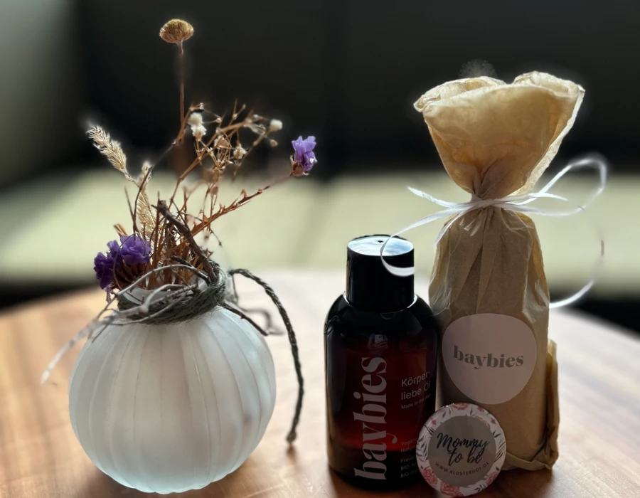 Dekorative Anordnung mit einer Glasvase, Trockenblumen, einem Körperöl und einem Geschenk für werdende Mütter auf einem Holztisch.