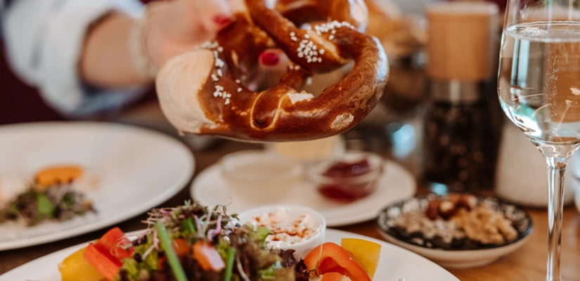 Eine Hand hält eine frisch gebackene Brezel über einem Tisch mit Salat und Getränken in einem gemütlichen Restaurant.