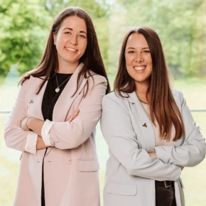 Two women smiling, standing side by side with arms crossed, wearing light-colored blazers in a bright, natural setting.