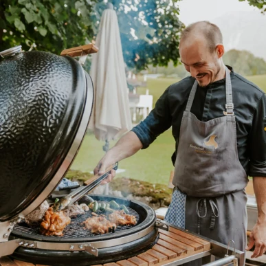 Ein Koch grillt Fleisch und Gem&uuml;se auf einem gro&szlig;en Grill im Freien, umgeben von einer sch&ouml;nen, gr&uuml;nen Landschaft.