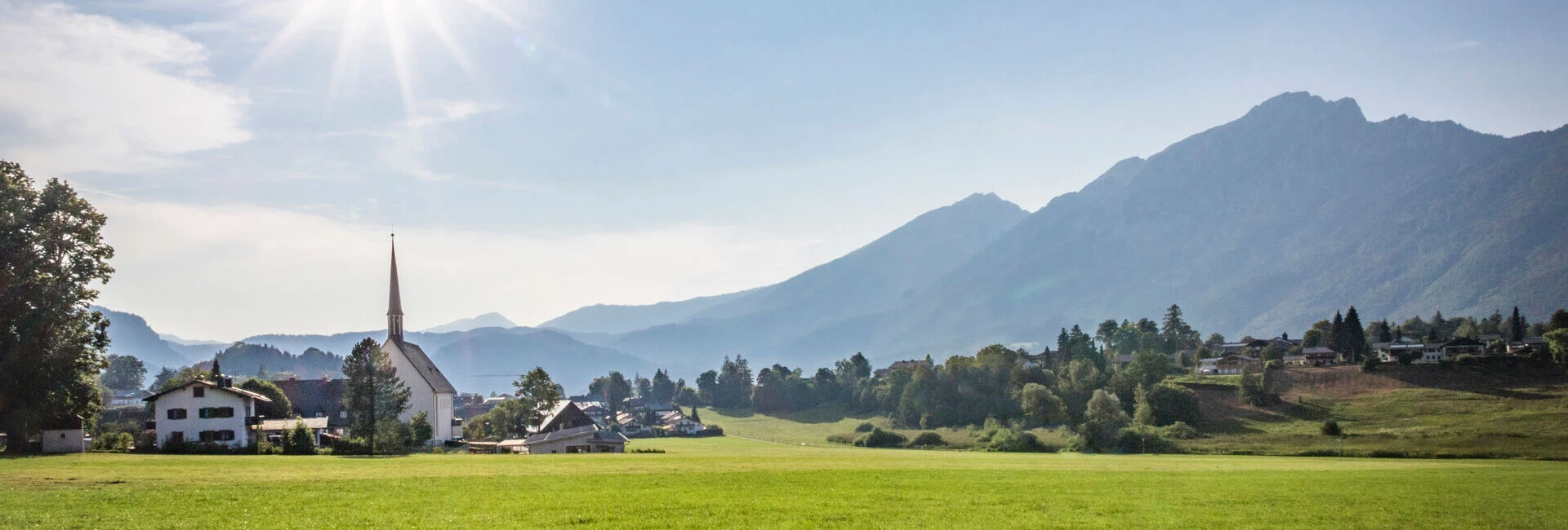 Sonniger Blick auf die Landschaft mit einer Kirche in Bayerisch Gmain, umgeben von Bergen und Wiesen.