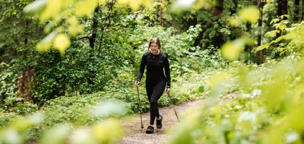 Eine Frau wandert mit St&ouml;cken auf einem Waldweg, umgeben von &uuml;ppigem Gr&uuml;n und B&auml;umen.