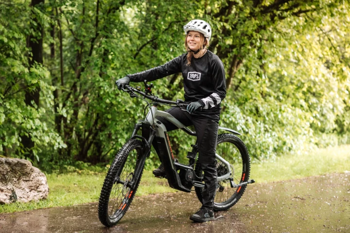 Eine Radfahrerin in Schutzkleidung posiert lächelnd auf ihrem E-Bike in einer grünen, bewaldeten Umgebung.