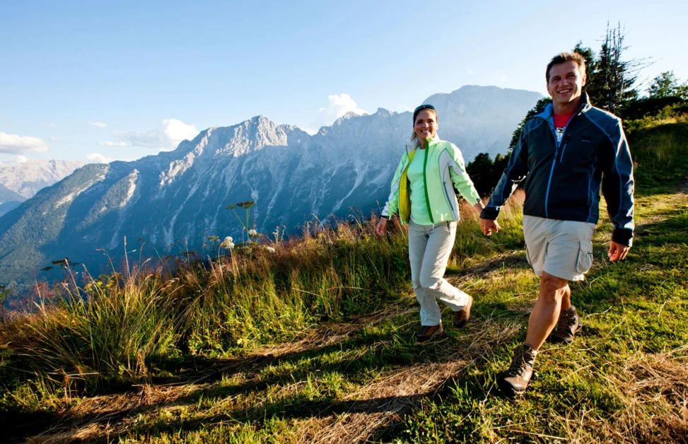A couple hikes on a grassy trail with scenic mountain views under a clear sky.