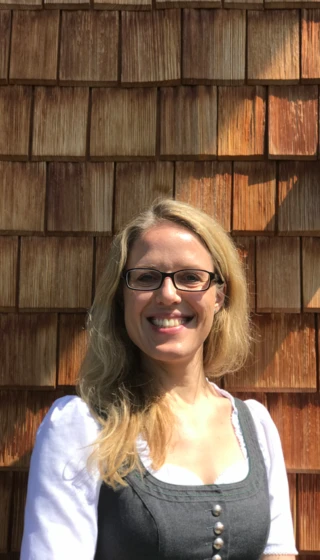 Eine lächelnde Frau in Tracht steht vor einer Holzschindelwand. Sonnenschein betont die freundliche Atmosphäre.