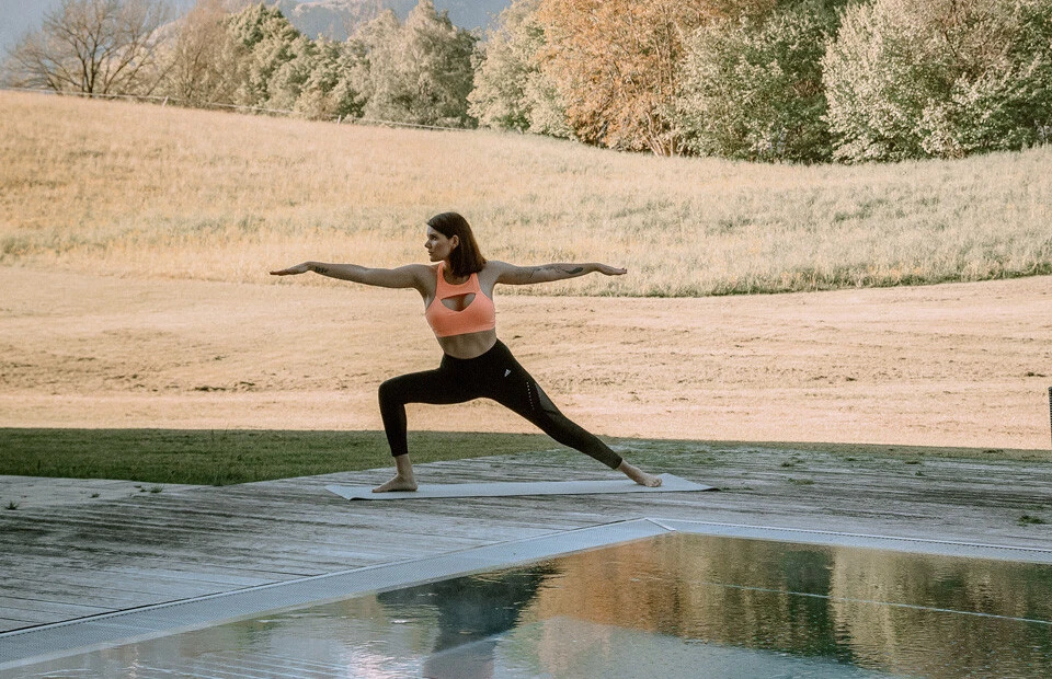 Eine Frau übt Yoga im Freien vor einer Berglandschaft, mit einem Pool im Vordergrund, umgeben von Natur in Bayern.