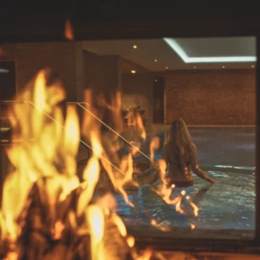 Blick durch ein Fenster auf einen Poolbereich mit einer Person im Wasser, umgeben von warmem Licht und einem Feuer im Vordergrund.