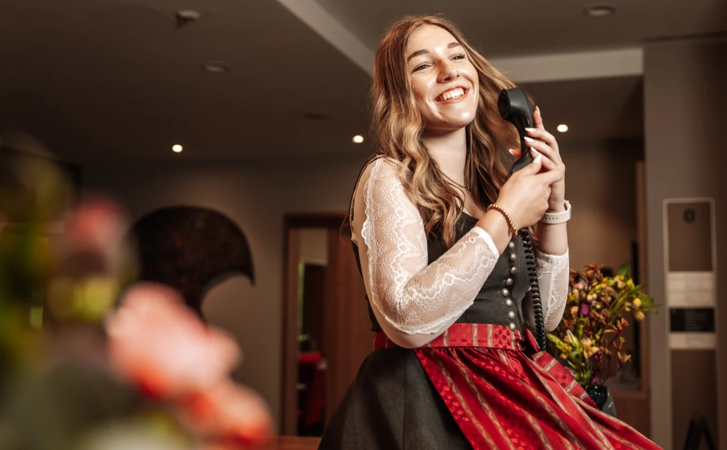 Eine lächelnde Frau im traditionellen Outfit spricht am Telefon in der Lobby des Hotel Klosterhof in Bayern.