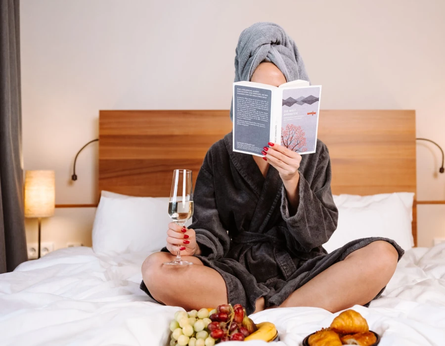 Eine Frau in einem Bademantel sitzt im Bett, liest ein Buch und genießt Sekt, umgeben von frischem Obst und Gebäck im Hotel Klosterhof.