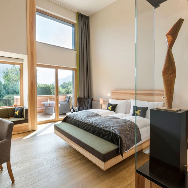 Modern hotel room with large windows offering mountain views, featuring a bed, desk, seating area, and balcony. Wellnesshotel Klosterhof in Bayern.