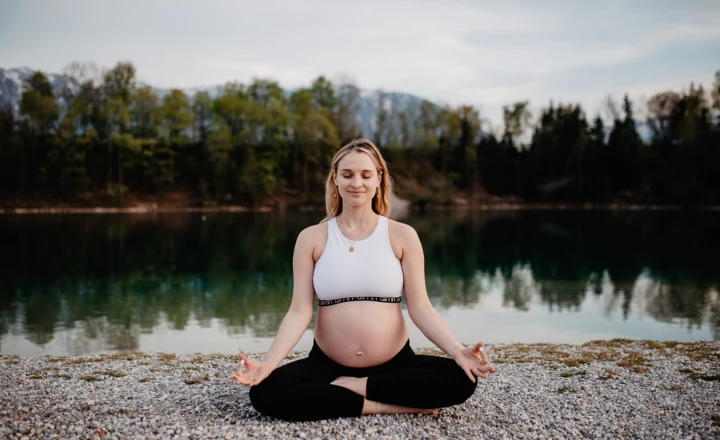Eine schwangere Frau meditiert am Ufer eines Sees vor einer bewaldeten Kulisse.
